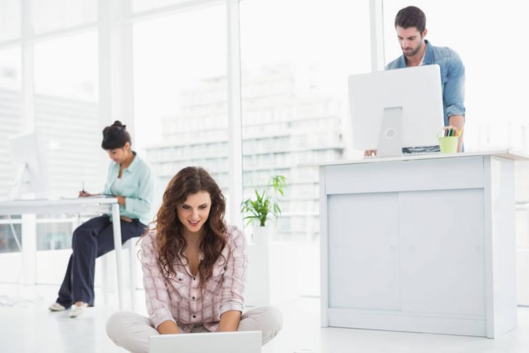 businesswoman sitting on the floor using laptop with colleagues standing behind