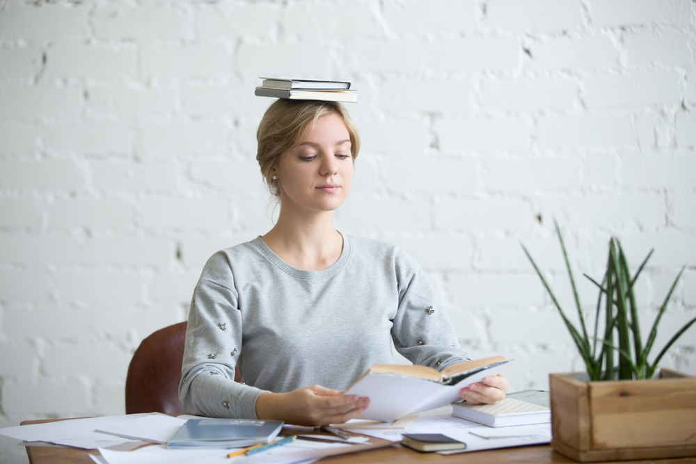 woman is sitting straight at the desk with books on her head