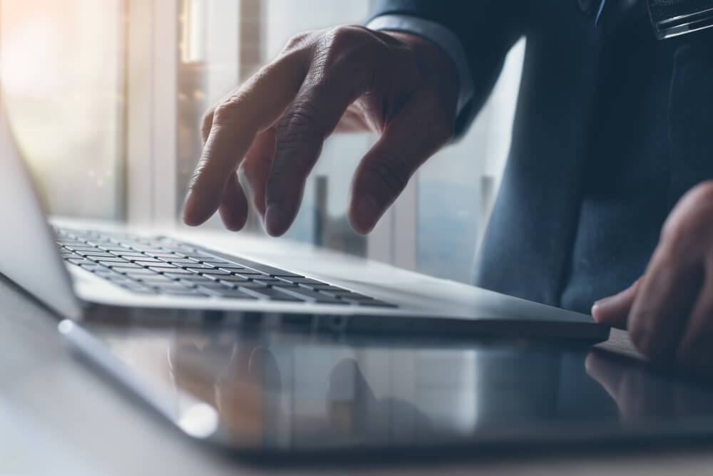 Businessman in black suit standing at the adjustable desk and typing on laptop