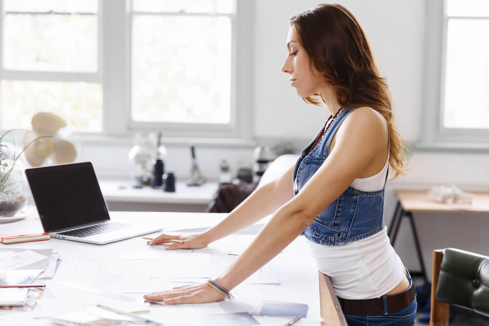 Young woman is standing at sit-stand desk in creative office